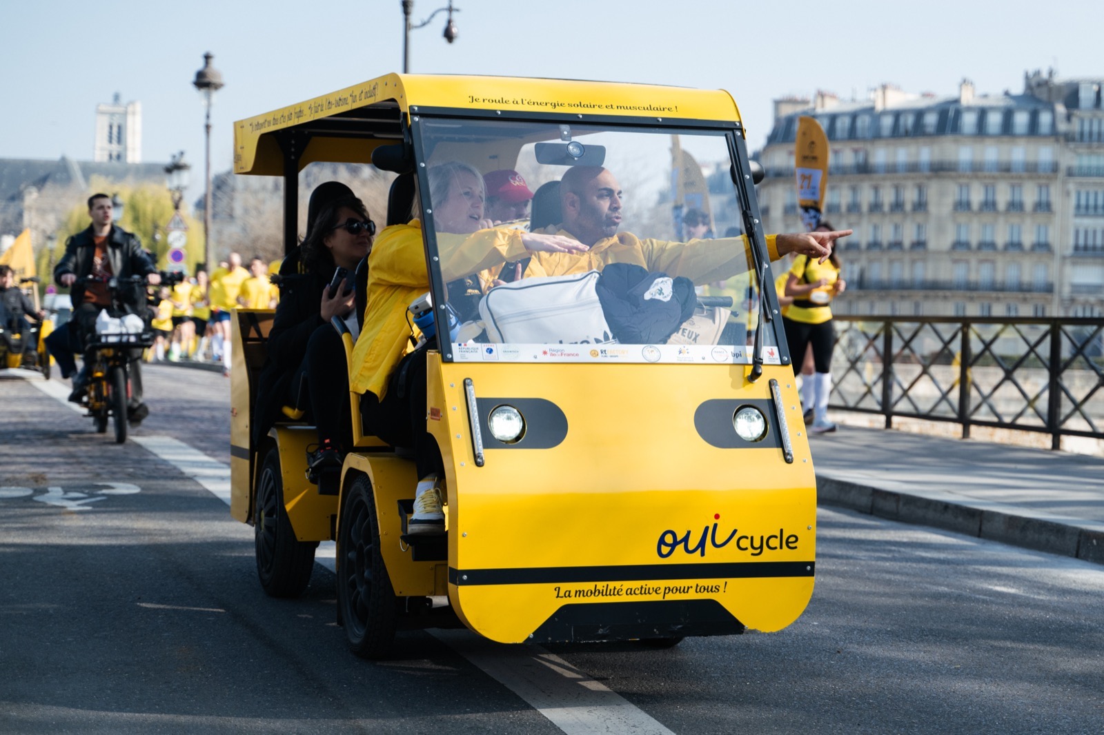 Le OuiCycle jaune sur un pont de Paris : le conducteur désigne un point d’intérêt à ses passagers.