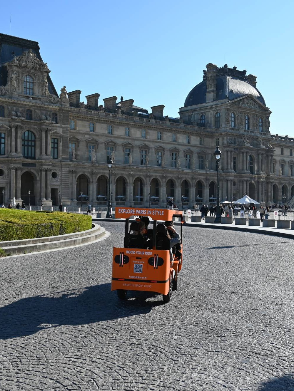 Un OuiCycle orange vu de l’arrière, chargé de visiteurs, tourne devant le Louvre.
