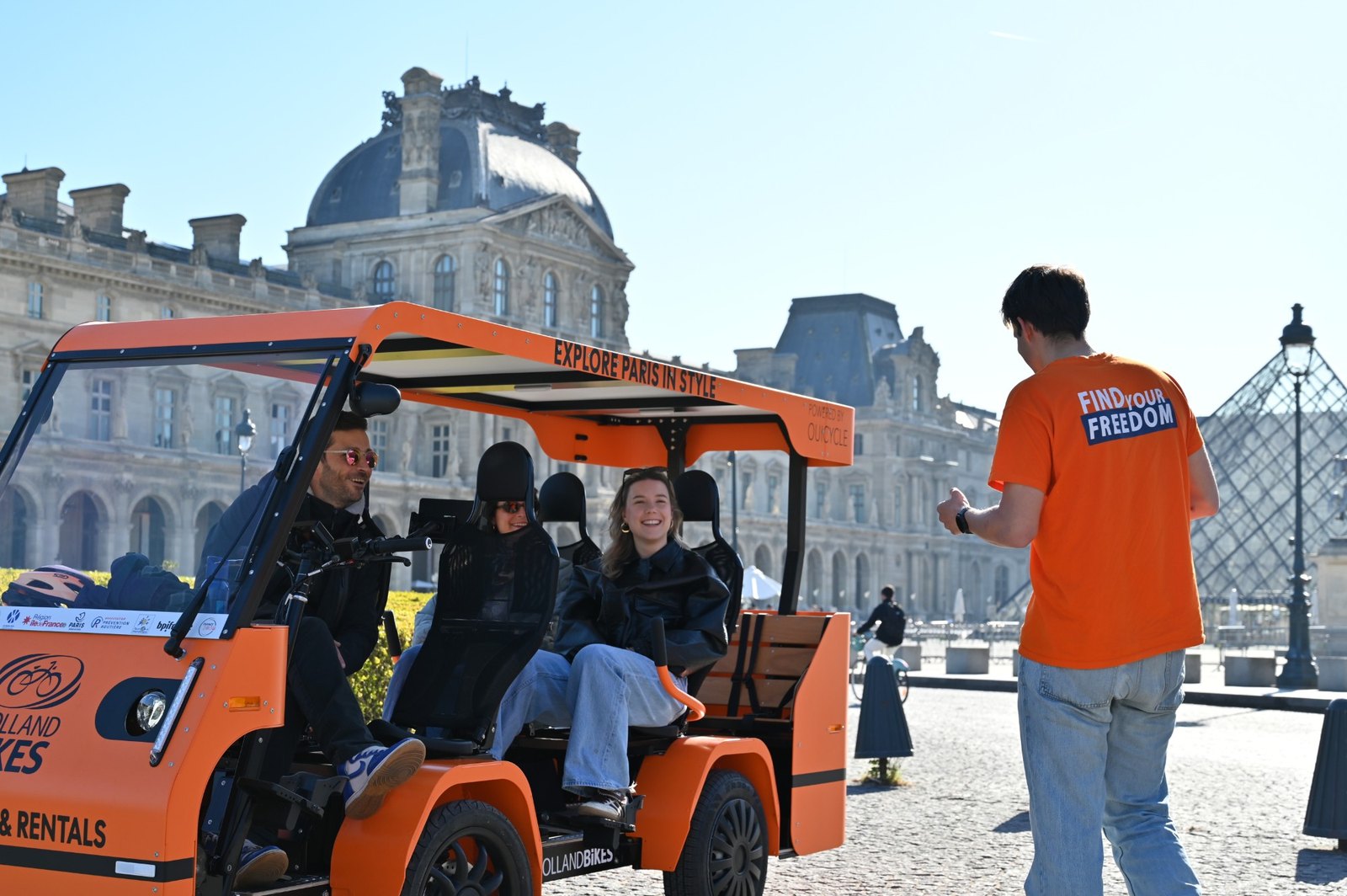 Des visiteurs rient à bord d’un OuiCycle orange devant le Louvre, accueillis par un guide.